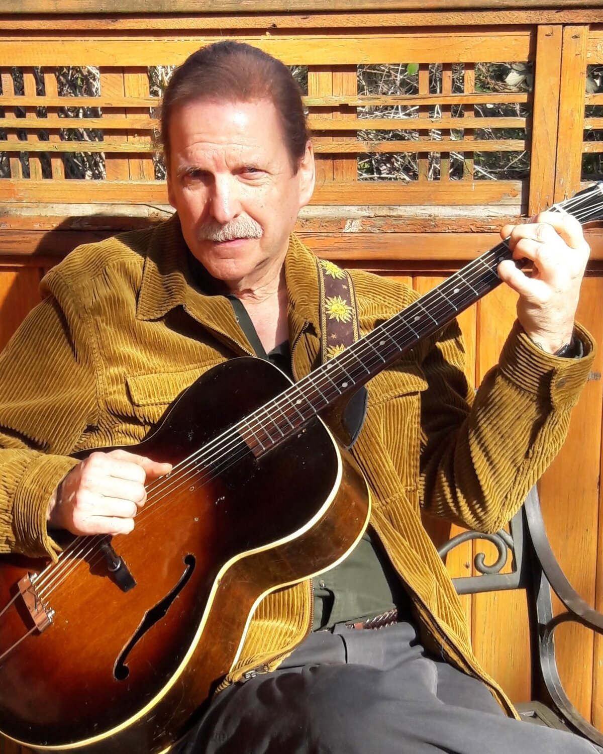 Dan Abernethy in a corduroy jacket, playing a sunburst archtop guitar in front of a cedar fence